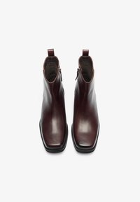 Botas de tobillo de cuero marrón con un acabado suave, punta cuadrada y cremalleras laterales. Cuentan con una pestaña en la parte trasera y una suela negra ligeramente elevada.