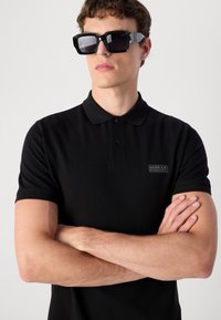 Polo noir en tissu texturé, avec un col à trois boutons et des manches courtes. Présente un petit patch logo sur la poitrine gauche.