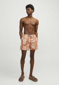 Zwemshorts in licht perzik met witte abstracte patronen, elastische tailleband, zijzakken en een trekkoord. Draag het met bruine slippers.