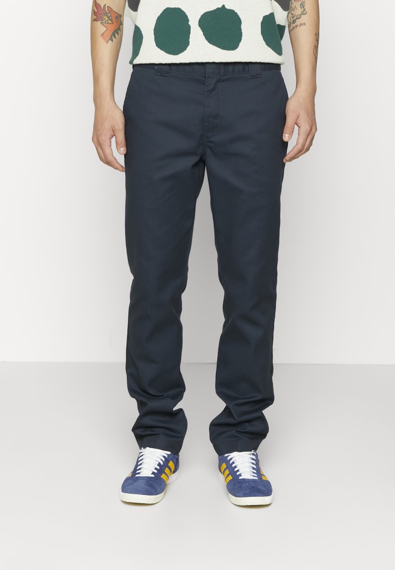 Homme portant un pantalon bleu marine foncé, des baskets bleues avec des rayures jaunes et une chemise blanche à gros pois verts, debout devant un fond blanc.