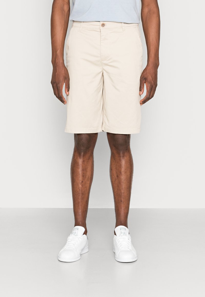 Pantaloni corti beige realizzati in tessuto leggero, dotati di chiusura con bottone, tasche laterali e una vestibilità dritta, abbinati a sneakers bianche.