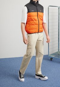 Puffa bez rukávů s černou horní částí a oranžovým spodkem, s předním zipem. Béžové cargo kalhoty s kapsami, doplněné černými teniskami s bílou podrážkou.