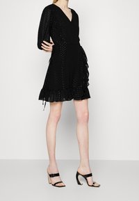 Svart omlottklänning i texturerad tyg, V-ringad halsringning, långa puffärmar och volangdetaljer i fållen. Bärs med svarta högklackade sandaler med remmar.