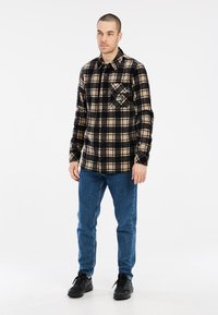 Camisa de franela a cuadros negra y beige con un bolsillo en el pecho, combinada con jeans azules y zapatos negros, llevada por una persona de pie.
