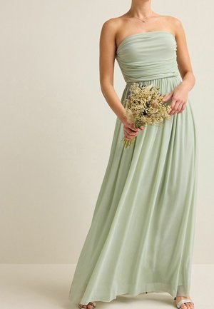 Femme en robe vert clair sans bretelles tenant un bouquet de fleurs séchées sur un fond beige uni.
