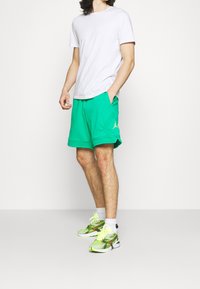 Bílý tričko s kulatým výstřihem, zelené sportovní šortky s bočními kapsami a detailním logem. Zelené a černé tenisky s vzorem a jasnými akcenty.