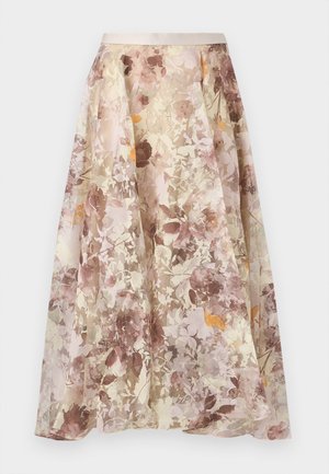 Femme portant un top sans manches violet foncé à volants, associé à une jupe fleurie asymétrique et des talons nude avec bride à la cheville, debout sur un fond uni.