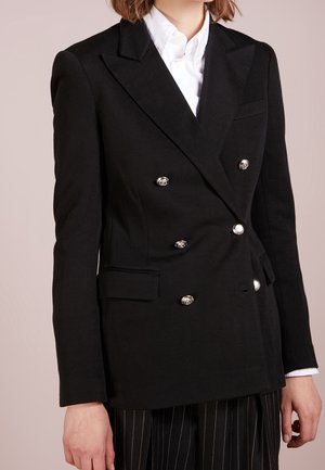Vrouw draagt een zwarte dubbelgeknoopte blazer met zilveren knopen over een wit overhemd en een zwarte pijpenkrant broek.