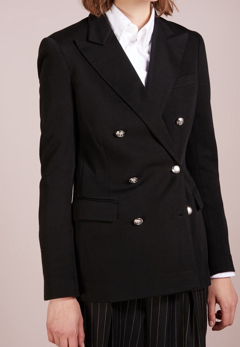 Femme portant un blazer noir à double boutonnage avec des boutons argentés sur une chemise blanche et un pantalon noir à fines rayures.