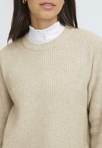 Béžový pletený sveter s textúrovaným vzorom; má okrúhly výstrih a je doplnený bielou volánkovou golierom, ktorý vyčnieva pod ním.