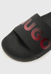 Μαύρο σανδάλι slide με υφασμένη επιφάνεια πέλματος και ευρύ λουράκι που διαθέτει μεγάλα κόκκινα γράμματα με διαβάθμιση, σχηματίζοντας μέρος ενός ονόματος μάρκας.