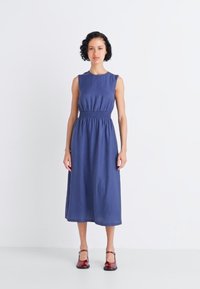 Blue, sleeveless dress with a smocked waist, midi length, and smooth texture. Paired with red shoes, standing against a plain background.