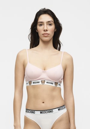 Młoda kobieta o ciemnych włosach nosi jasnoróżowy biustonosz z logo misiów oraz białe majtki z nazwą marki Moschino na gumce.