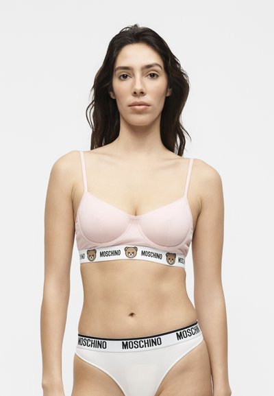 Jeune femme aux cheveux foncés porte un soutien-gorge rose clair avec des logos d’ours et un sous-vêtement blanc avec le nom de la marque Moschino sur la ceinture.