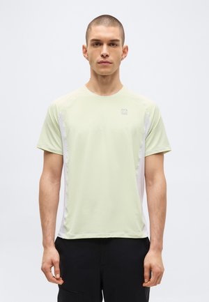 Jeune homme aux cheveux courts face à l’avant, portant un t-shirt de sport vert pâle à manches courtes et un pantalon noir sur un fond uni.