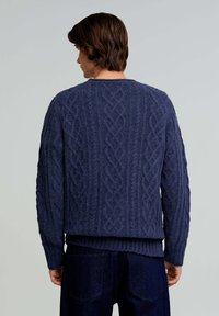 Marineblaue Strickjacke mit strukturiertem Kabelmuster, gerippten Bündchen und Saum. Über dunklen Jeanshosen getragen, Blick von hinten.