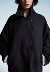 Črn volnen plašč z visokim ovratnikom, dvema velikima gumboma in žepi podloženimi s kremasto tkanino. Mehka tekstura in prevelik dizajn.