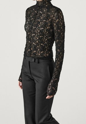 Weibliches Model trägt einen transparenten schwarzen floralen Spitzen-Longsleeve-Oberteil mit hohem Kragen und schwarze maßgeschneiderte Hose, steht vor einem einfarbigen Hintergrund.