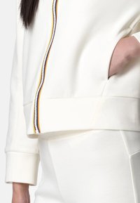 Felpa bianca con zip, dotata di una cerniera a righe colorate, polsini a costine e tasche laterali. Materiale morbido con un design minimalista.