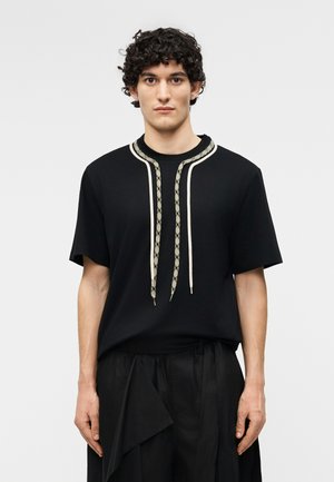 Jeune homme aux cheveux bouclés portant une chemise noire à manches courtes avec des cordons à motifs et un pantalon noir ample sur un fond blanc uni.