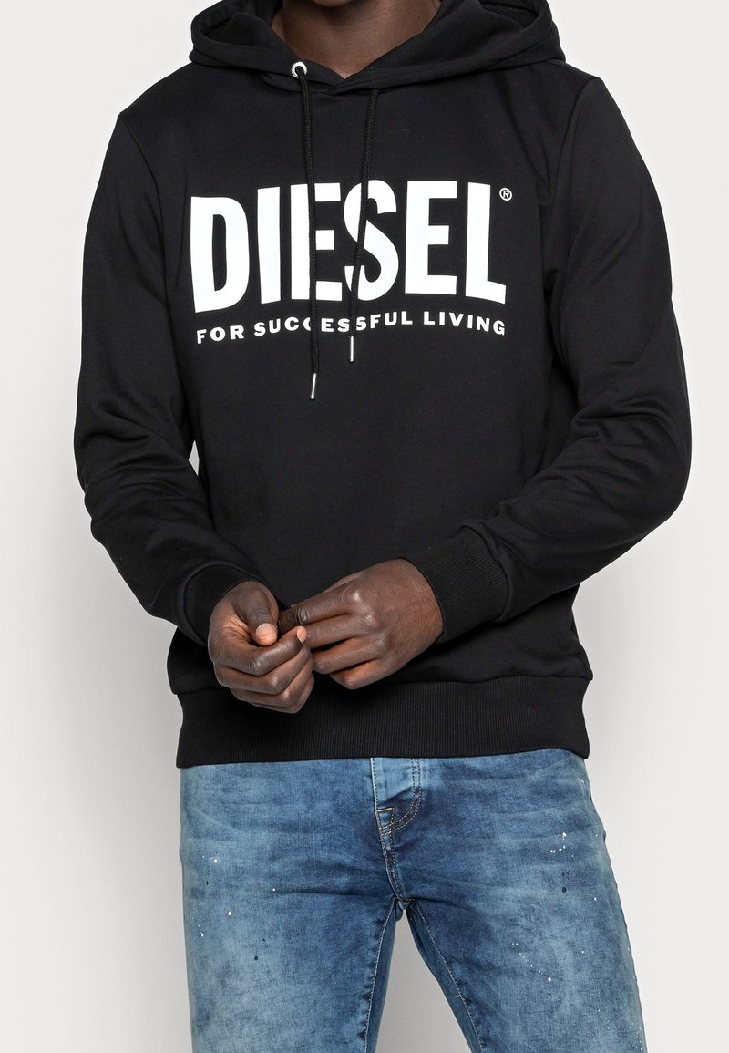 Hoodie noir avec le texte blanc "DIESEL FOR SUCCESSFUL LIVING", porté par une personne aux mains jointes, associé à un jean bleu.