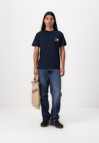 T-shirt en coton bleu marine avec un petit graphique sur la poitrine gauche, associé à un jean bleu et un sac beige. Le modèle a les cheveux longs.
