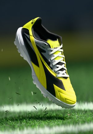 Soccer cleat with a yellow and black design, textured upper, white laces, and molded studs on a white outsole, suspended above grass.