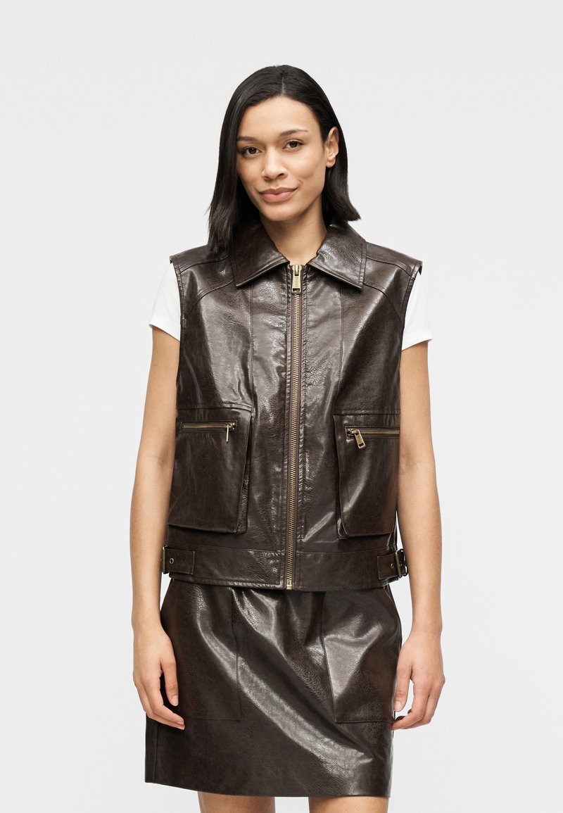 Καφέ δερμάτινο γιλέκο με φερμουάρ μπροστά, μεγάλο γιακά και δύο τσέπες στο στήθος. Το υλικό έχει υφή με γυαλιστερό φινίρισμα.