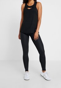 Svart ärmlös träningstopp med utskärningsdetaljer, parat med svarta leggings och vita träningsskor på en neutral bakgrund.