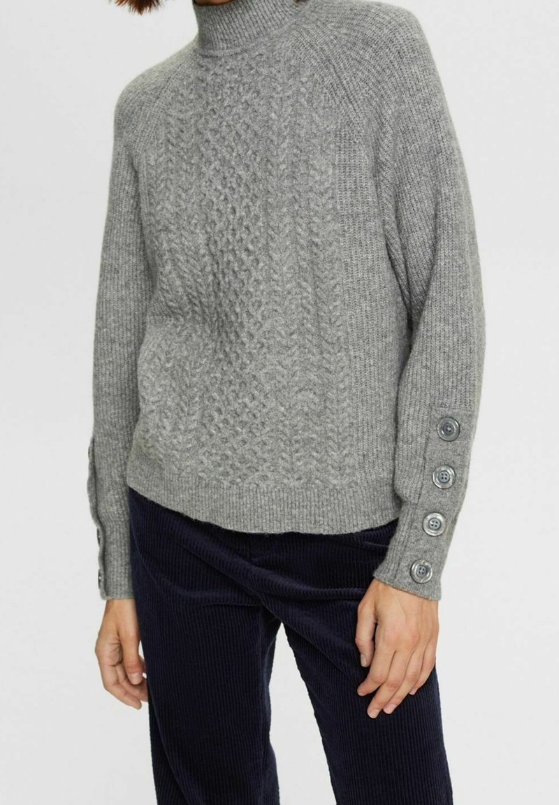 Pull en maille grise avec un col montant, présentant un motif en torsades et des accents de boutons aux poignets. Texture douce et coupe décontractée.