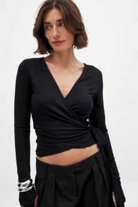 Schwarzes, langärmeliges Wickeloberteil aus weichem Material mit V-Ausschnitt und gerafftem Detail. Kombiniert mit hoch taillierten schwarzen Hosen und silbernen Armbändern.