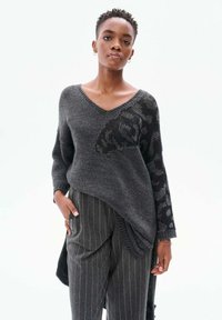 Grauer Strickpullover mit V-Ausschnitt, der ein strukturiertes schwarzes Muster an einem Ärmel aufweist und einen längeren Rückensaum hat. Kombiniert mit grauen Nadelstreifen-Hosen.