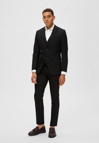 Schwarzer Anzug mit tailliertem Blazer, passender Hose und weißem Hemd. Braune strukturierte Schuhe mit Quasten. Glattes Material und maßgeschneiderte Passform.