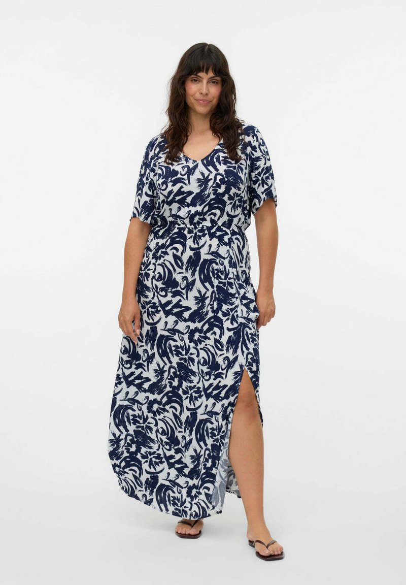 Donna con capelli scuri che indossa un maxi abito floreale blu navy e bianco con uno spacco laterale, abbinato a sandali flip-flop neri, in piedi su uno sfondo bianco.