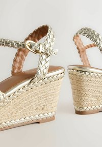 Sandales compensées tressées avec des lanières dorées tressées et une boucle à la cheville, avec des talons compensés enveloppés de jute sur un fond clair.