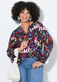 Multikleurige blouse met gedurfde "PEACE" tekst en hartpatronen, lange mouwen, lichtgewicht stof, gecombineerd met lichtblauwe jeans en een witte tas.