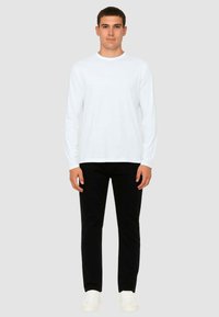 T-shirt bianca a maniche lunghe con scollo rotondo, abbinata a pantaloni neri aderenti e sneakers bianche. Il modello è in piedi su uno sfondo grigio.