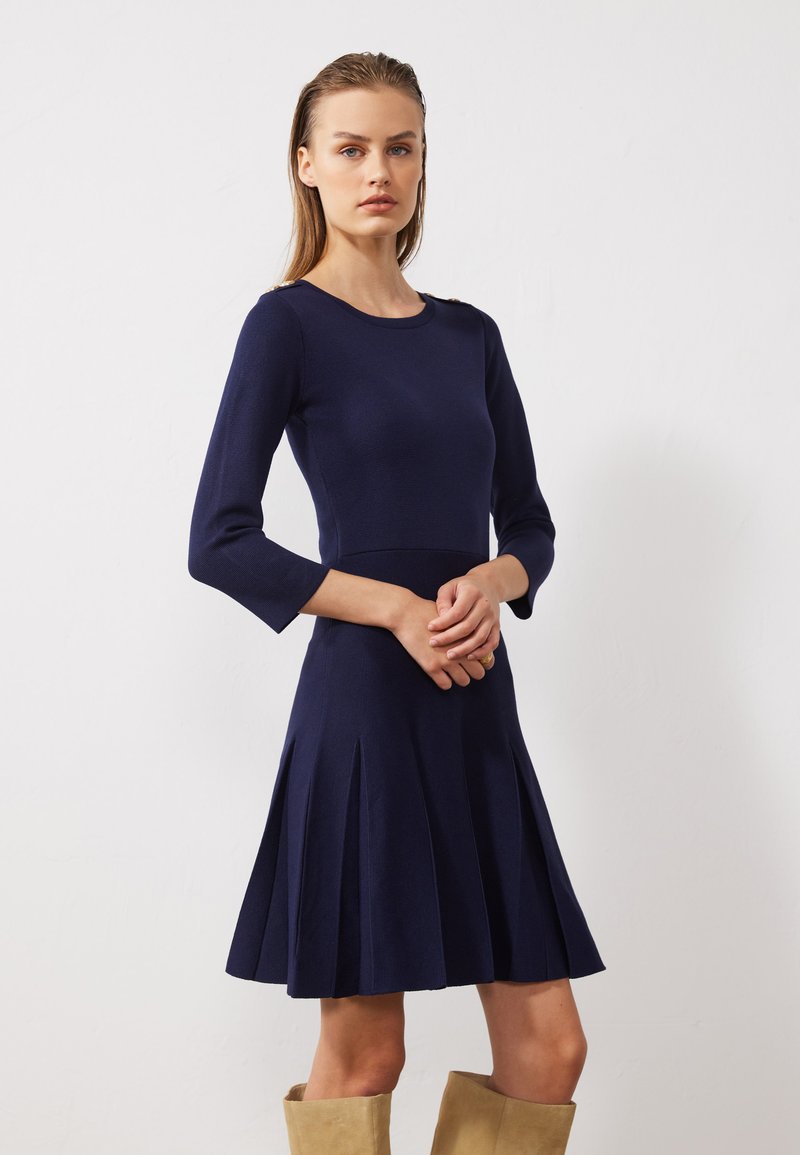 Robe bleu marine avec des manches 3/4, un corsage ajusté, une jupe évasée et des plis. Matière tricotée avec un col rond et des détails subtils aux épaules.