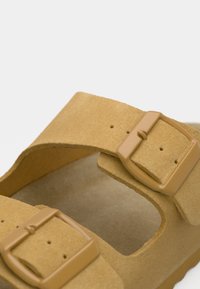 Ταμπά σανδάλι με δύο ρυθμιζόμενους ιμάντες με πλαστικές αγκράφες, κατασκευασμένο από μαλακό υλικό που μοιάζει με σουέτ. Η εσωτερική σόλα φαίνεται να έχει επένδυση για άνεση.