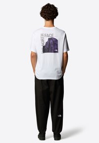 Camiseta de algodón blanca con un gráfico morado de una montaña y el texto "THE NORTH FACE" en la parte posterior. Combinada con pantalones y calzado negros.