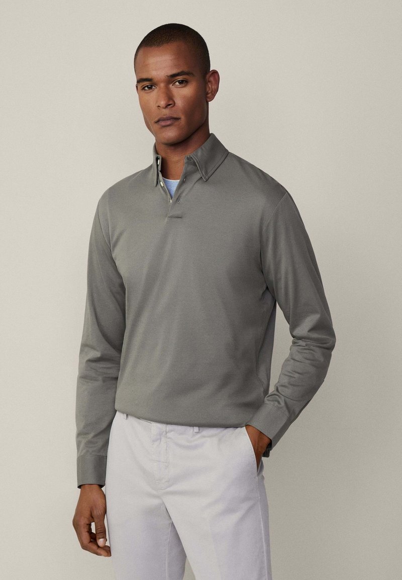 Siva dugmadna polo majica s dugim rukavima i ovratnikom, sa suptilnom plaketom i glatkom teksturom, u kombinaciji sa svijetlim hlačama.