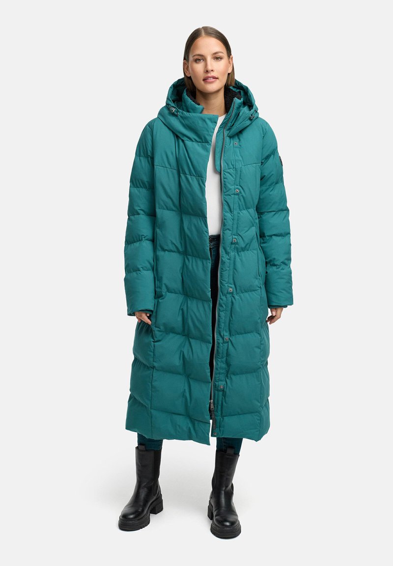 Tirkīza krāsa pūkains mētelis ar kapuci, kas tajā iekļauj quilted dizainu, priekšējus pogas un garas piedurknes. Vilkts virs baltas blūzes un melnām biksēm.