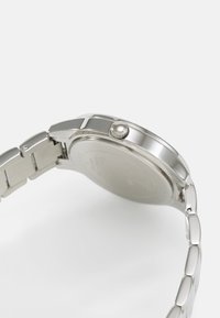 Montre en acier inoxydable argenté avec un boîtier rond, un bracelet maillé et une couronne texturée sur le côté. Fini lisse avec un design minimaliste.