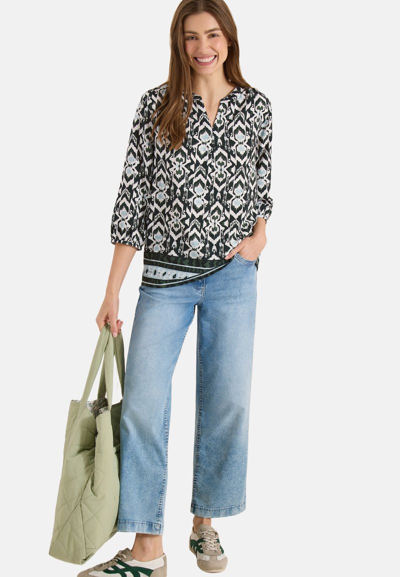 Femme souriante portant un chemisier à motifs noir et blanc, un jean large bleu clair, des baskets, et tenant un sac cabas matelassé vert clair.