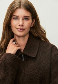 Braune, strukturierte Jacke mit Kragen und Reißverschluss. Das Model trägt einen silbernen Ring und posiert mit der Hand am Hals. Natürlicher Make-up-Look und lose Haare.