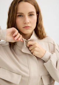 Chaqueta cremosa con cremallera, cuello alto y dos bolsillos frontales, tela suave, detalles visibles de cremallera y un ajuste holgado.