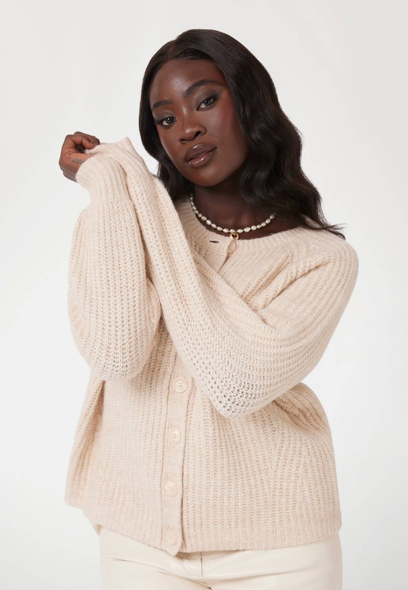 Beige Strickjacke mit übergroßen Ärmeln, vorne mit Knöpfen zu schließen und ribbeliger Textur. �Über hellfarbenen Hosen getragen.