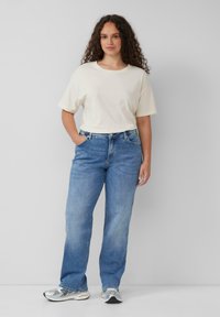 T-shirt en coton beige clair à manches courtes, associé à un jean en denim bleu clair. Modèle portant des chaussures de sport argentées. Design simple, coupe décontractée.