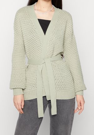 Cardigan en maille texturée vert clair à manches longues, larges poignets côtelés et ceinture assortie nouée à la taille, porté sur un top noir.
