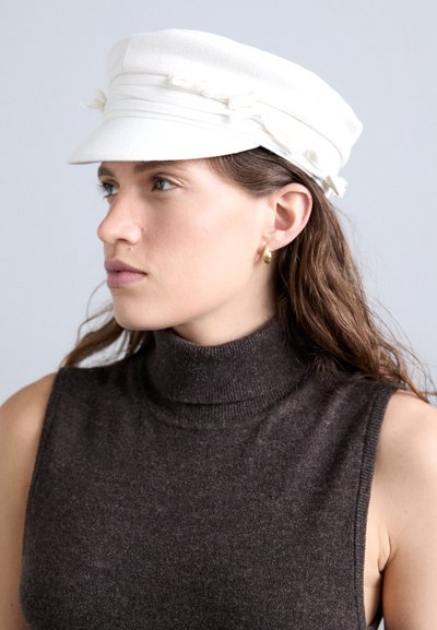 Cappello bianco con un design strutturato, dotato di visiera frontale e inserti in tessuto. Il modello indossa un dolcevita senza maniche di colore scuro.